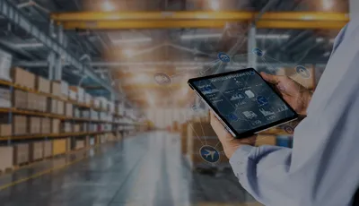 Pessoa a utilizar um tablet com interface digital sobreposta, num armazém moderno cheio de prateleiras com caixas, simbolizando a gestão digital da cadeia de abastecimento.
