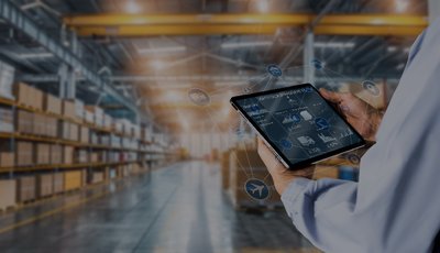 Pessoa a utilizar um tablet com interface digital sobreposta, num armazém moderno cheio de prateleiras com caixas, simbolizando a gestão digital da cadeia de abastecimento.