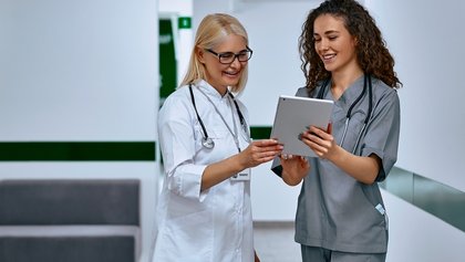 Duas profissionais de saúde a sorrir e a consultar um tablet num corredor de hospital.