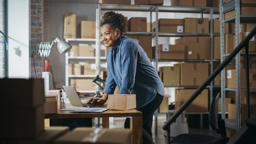 Mulher sorridente num armazém, a trabalhar com um computador portátil sobre uma secretária rodeada de caixas de cartão, representando gestão de inventário ou negócio online.