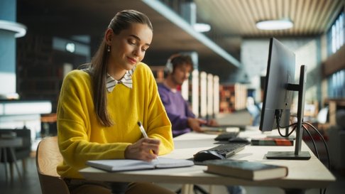 Estudante a estudar numa biblioteca moderna, sentada à secretária com um caderno e caneta, sorridente e concentrada, com computador e livros à sua frente