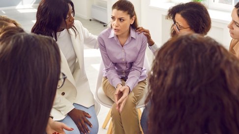 Grupo de mulheres sentadas em círculo, enquanto uma participante expressa emoções e recebe apoio empático das restantes, num ambiente que sugere uma sessão de partilha ou grupo de apoio emocional.