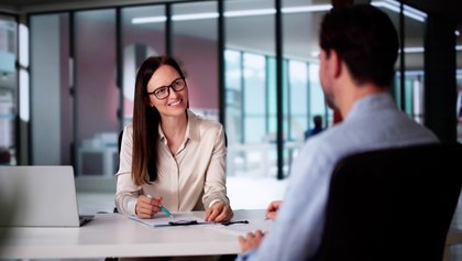 Mulher com óculos a sorrir e a escrever numa folha, durante uma conversa ou reunião de trabalho.